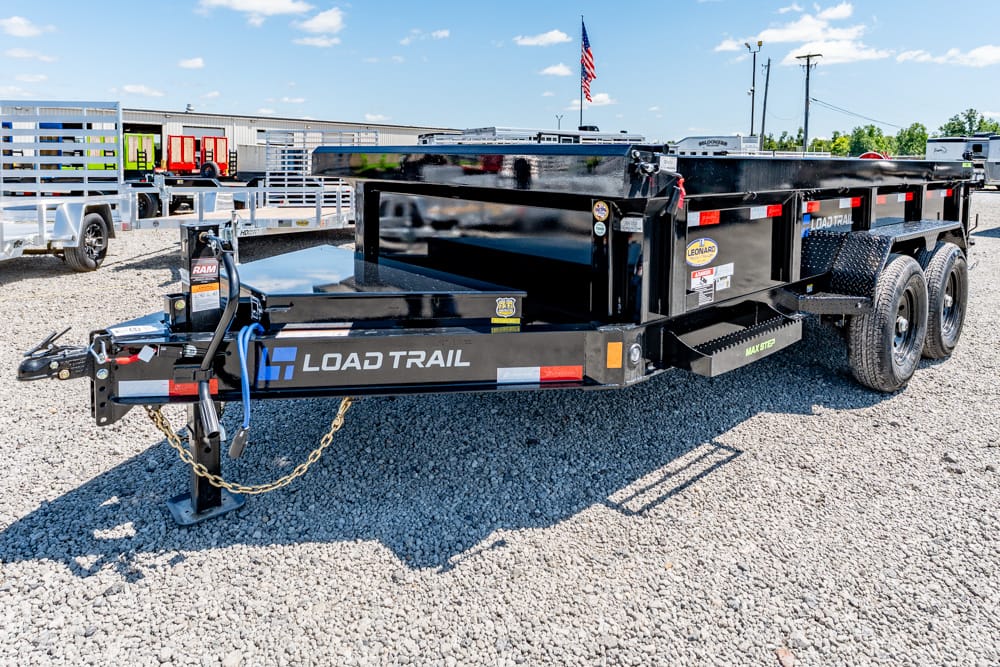 Black Load Trail tandem-axle dump trailer on a gravel lot with hitch, safety chains, and a blue sky background.