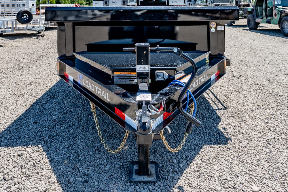 Front view of a Load Trail utility trailer on a gravel lot, showing hitch, safety chains, and a blue power cable.