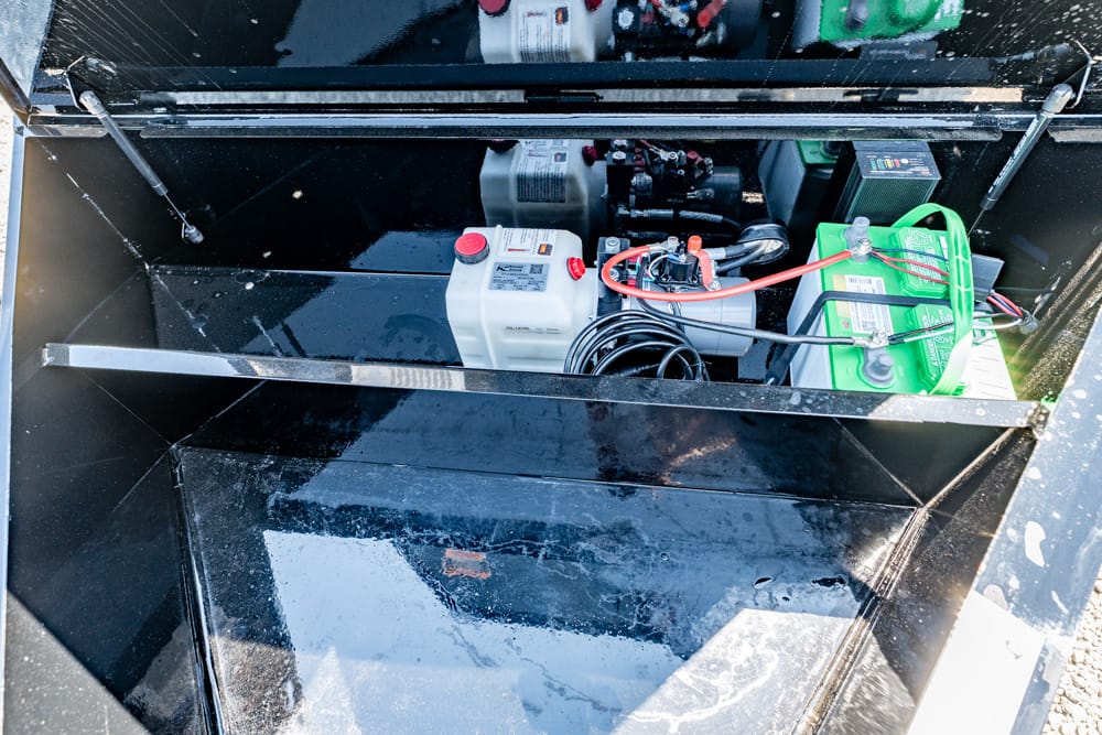 Open maintenance compartment with a green 12V battery, white fluid reservoir, and tangled cables around a small engine inside a glossy black housing.