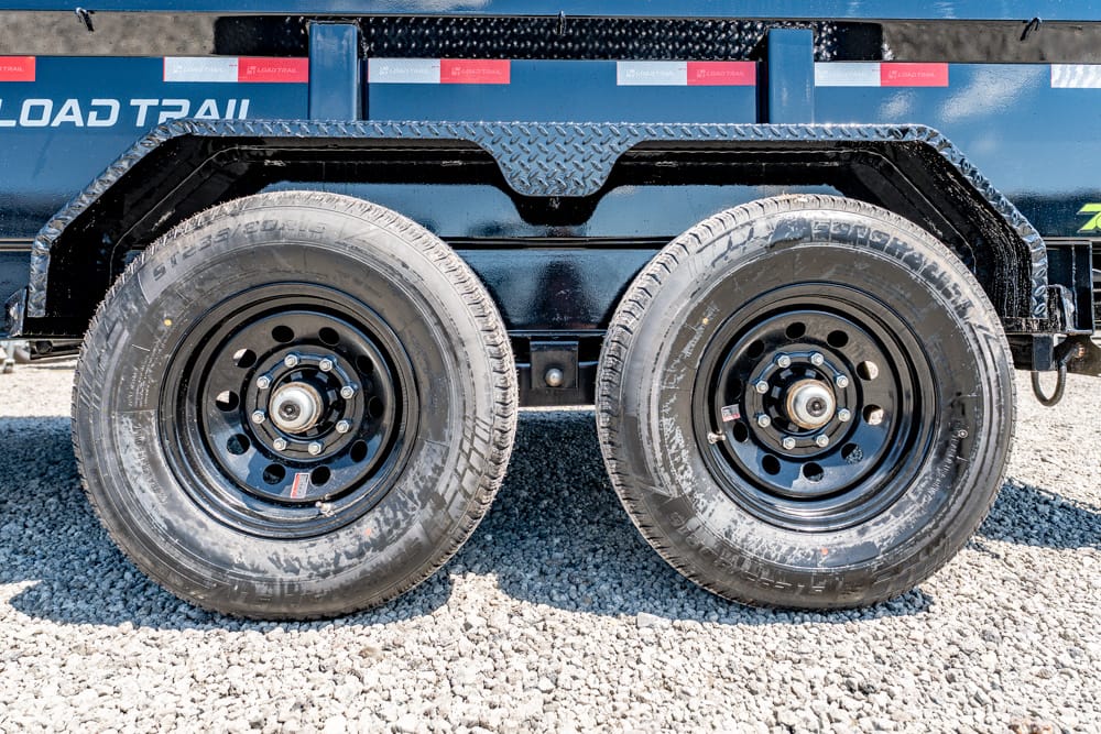 Tandem-axle trailer with two black wheels on a gravel surface; blue trailer body with 'LOAD TRAIL' text in the background.