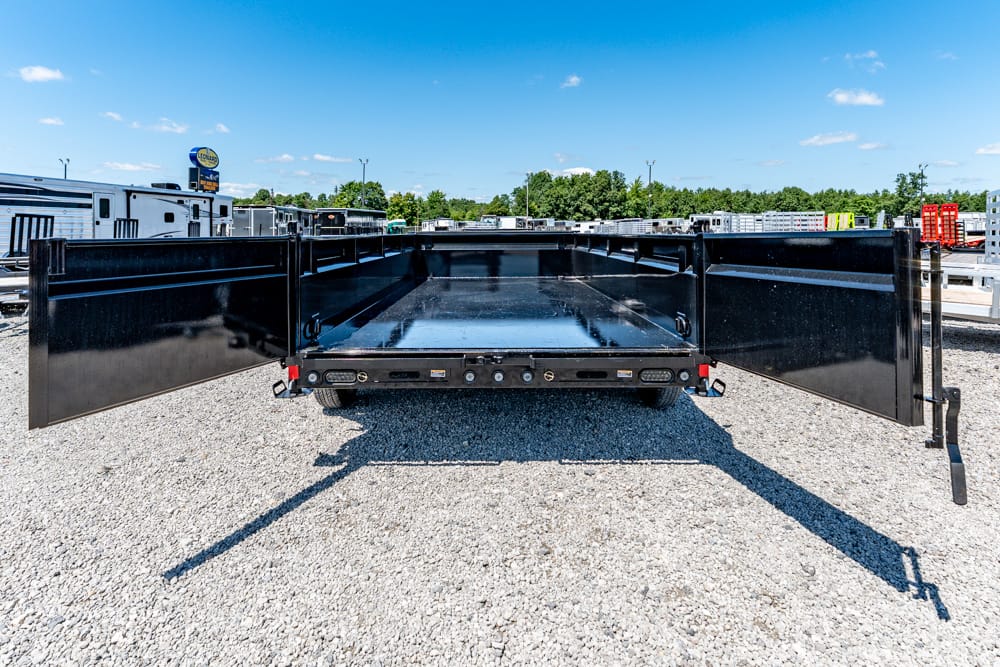 Rear view of a black flatbed trailer with its large side doors swung open, parked on a gravel lot under a blue sky.
