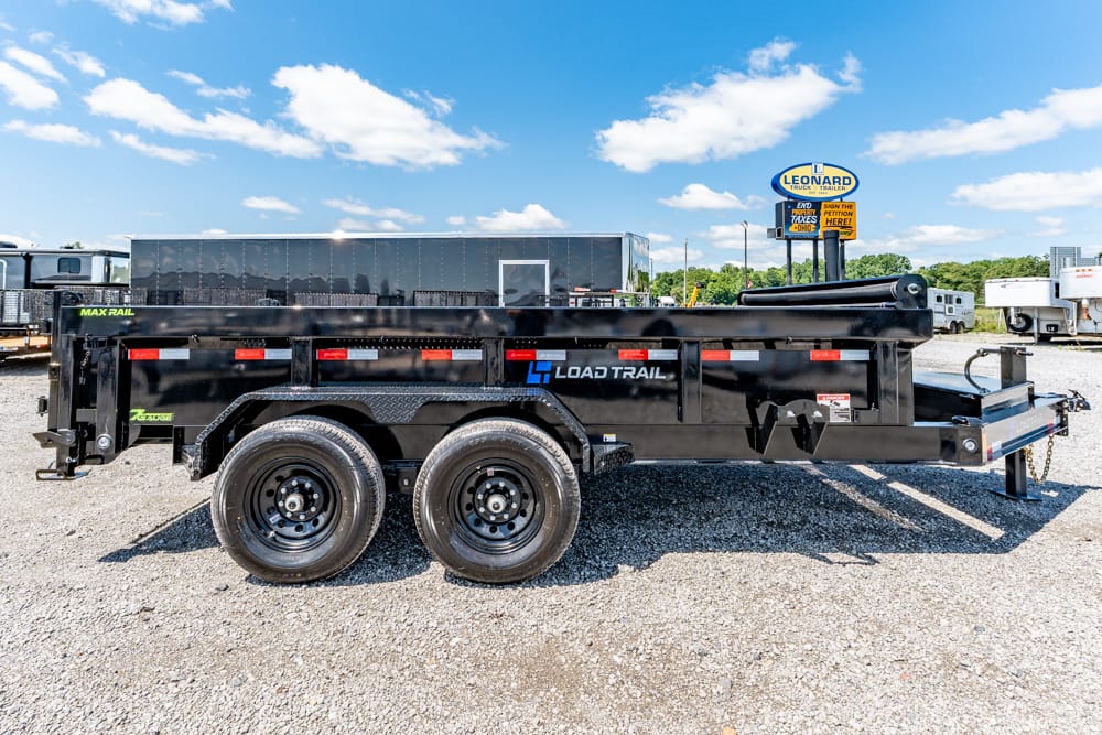 Black Load Trail tandem-axle trailer with a long steel bed, parked on gravel under a blue sky.
