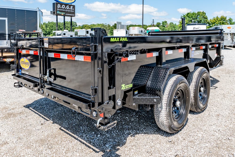 Black dual‑axle dump trailer with diamond‑plate fenders and MAX RAIL branding on a gravel lot at a trailer dealership.
