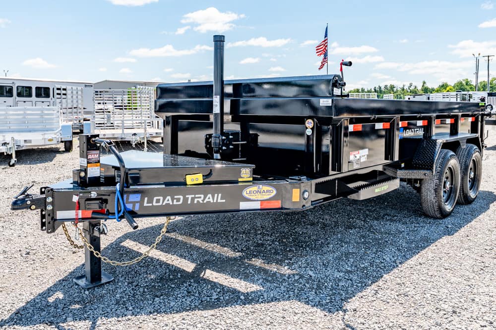 Black Load Trail dump trailer with dual axles and gooseneck hitch on a gravel lot; American flag in background