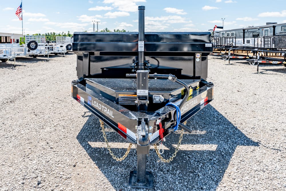 Front view of a black Load Trail trailer on a gravel lot, with hitch chains and a blue cord, other trailers in the background.
