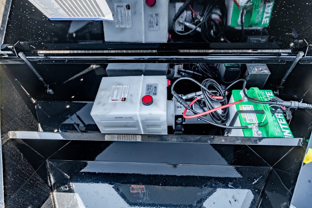 Open equipment compartment showing a white plastic reservoir with a red cap and a car-style battery with red and black cables, plus tangled wires inside a glossy black enclosure.