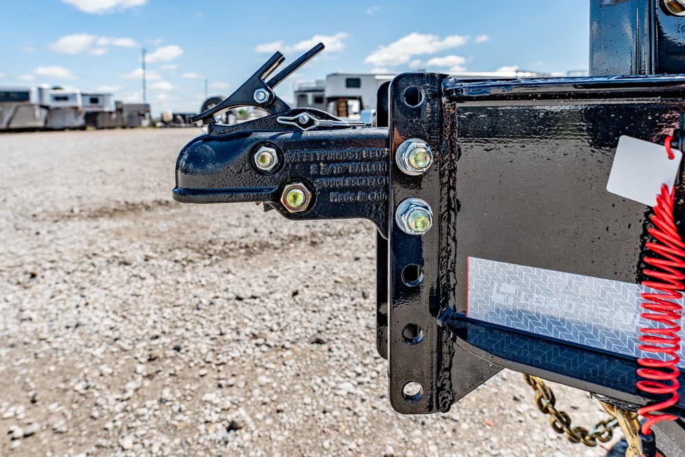 Close-up of a black trailer hitch with a latch, bolts, and a red coiled safety cord on a gravel lot.