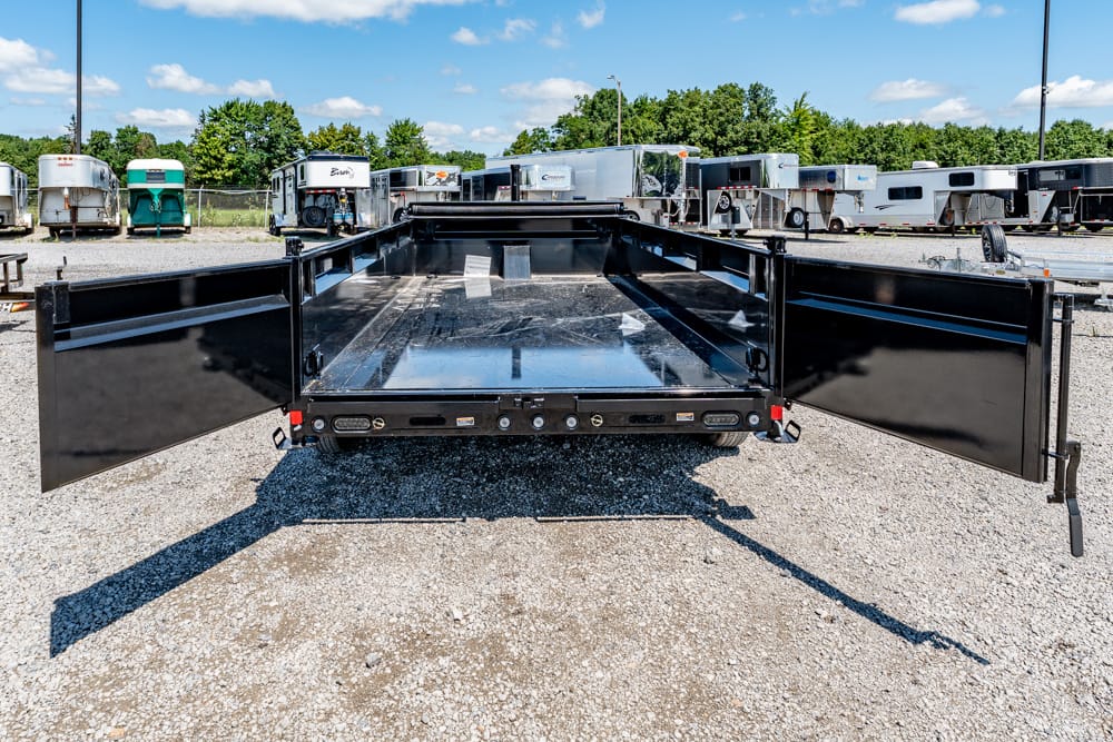 Back view of a black dump trailer with rear doors wide open, exposing an empty metal bed in a gravel trailer yard under a blue sky.