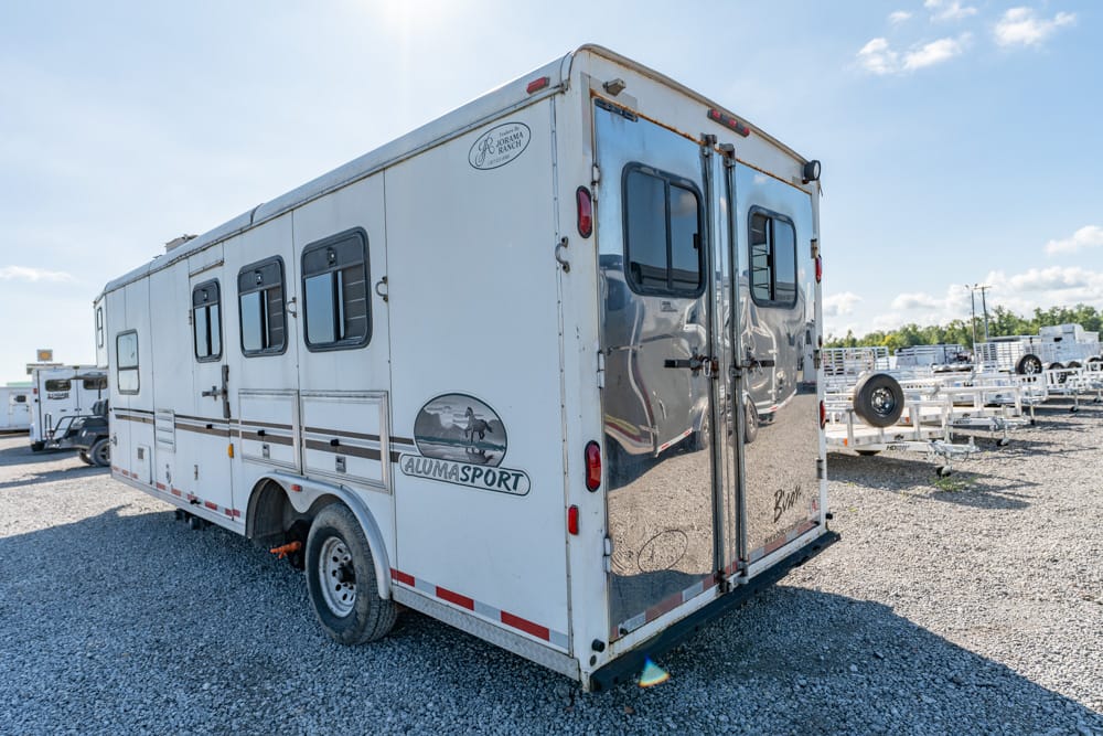 White horse trailer with AlumaSport logo parked on a gravel lot with other trailers in the background.