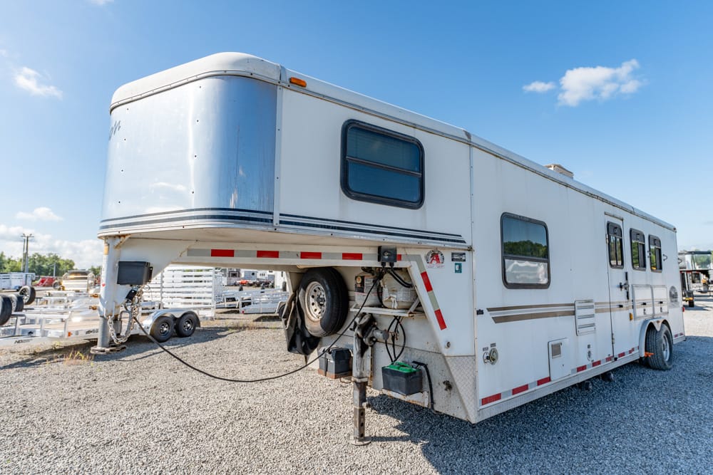 Large white gooseneck recreational trailer parked on a gravel lot under a clear blue sky, with spare tire and equipment mounted at the hitch area