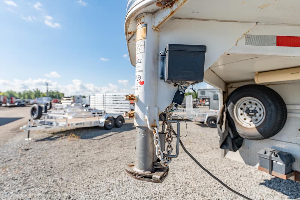 Close-up of a trailer hitch on a white utility trailer with a spare tire and safety chains, in a gravel yard.