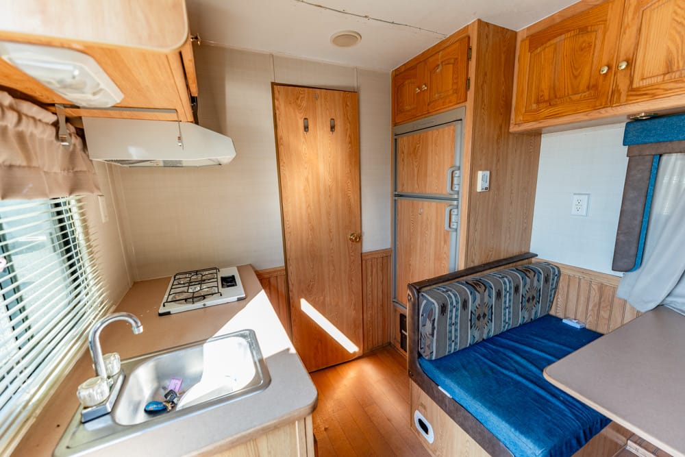 Compact RV kitchen with sink, stovetop, window blinds, and wooden cabinets beside a blue upholstered dinette.