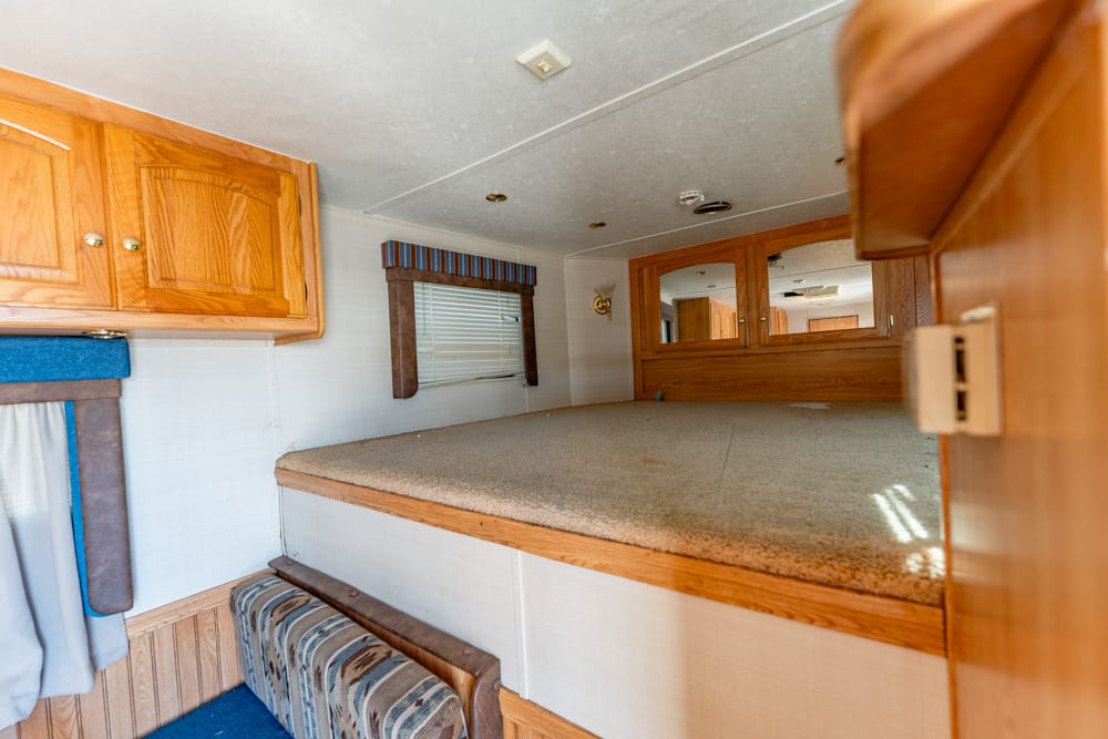 Compact RV interior showing an elevated sleeping platform with carpet, surrounding wood cabinets, and a small window with a striped valance around the corner