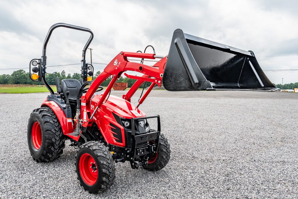 Red compact utility tractor with a front-end loader bucket on a gravel lot under a cloudy sky.
