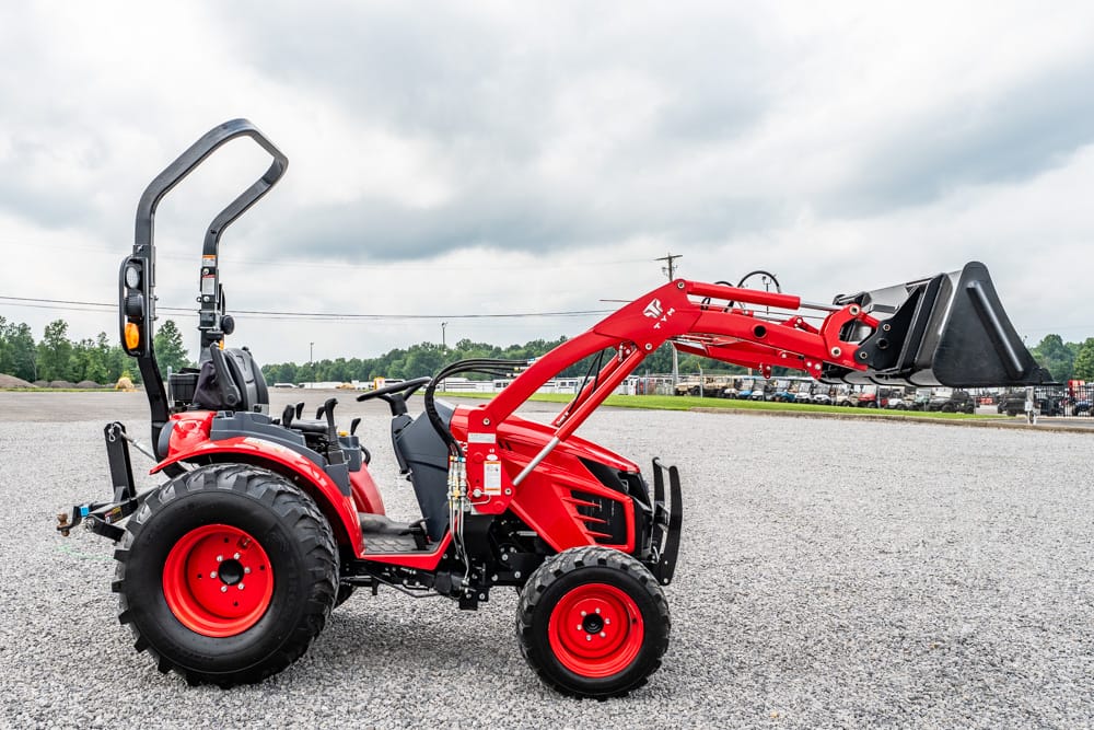 Red compact tractor with a front-end loader extending to the right, parked on a gravel lot under a cloudy sky.