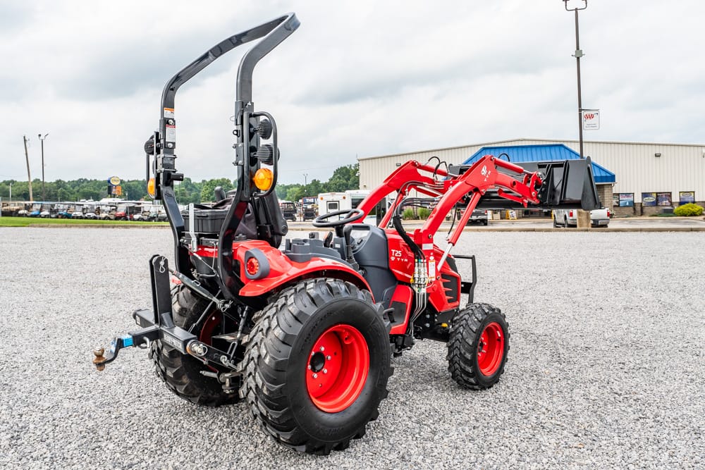 Red compact utility tractor with a front loader attachment on a gravel lot, facing right, with a building in the background.