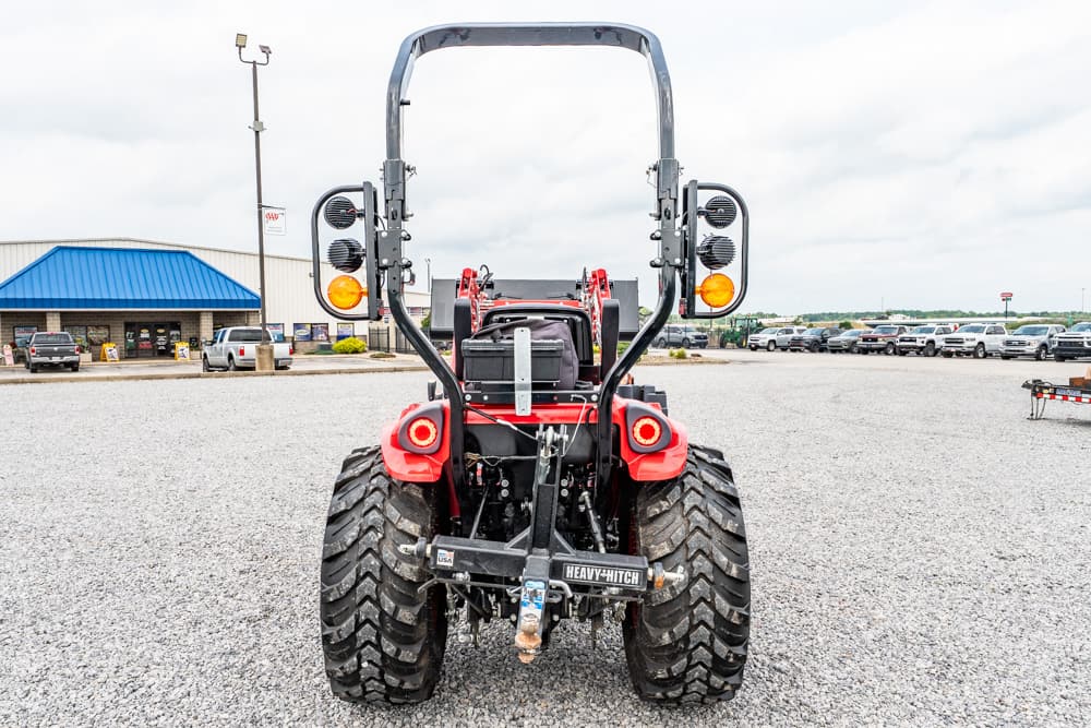Rear view of a red tractor with oversized off-road tires, roll bar, and hitch in a gravel parking lot.