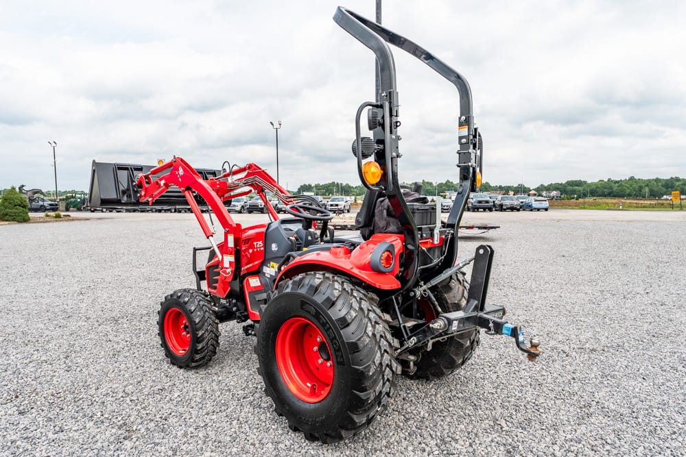Red compact tractor with a front loader and backhoe attachment parked on a gravel lot beside a row of cars.