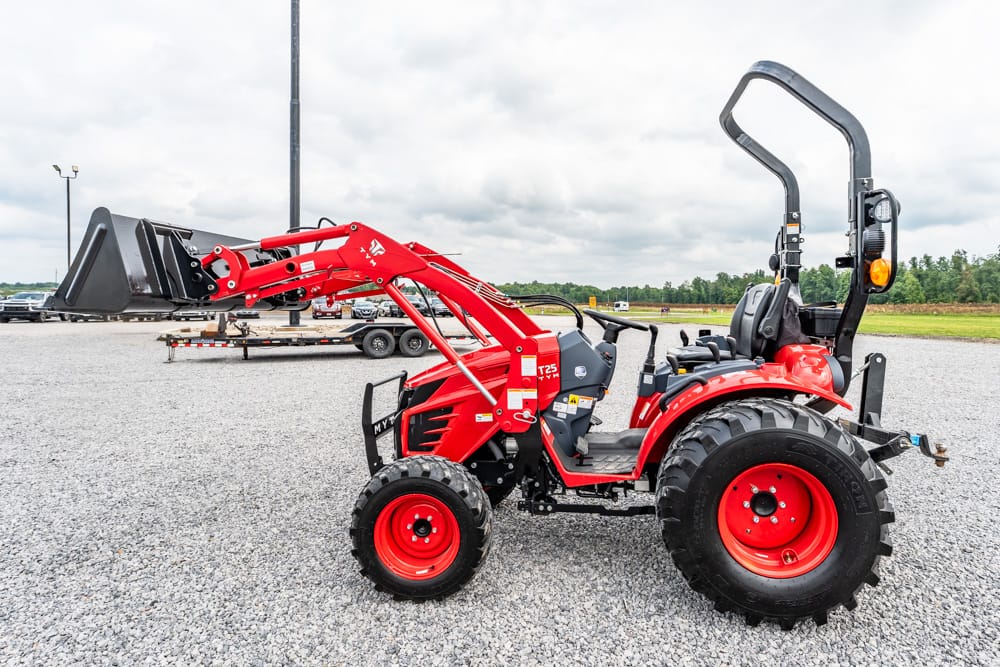 Red compact tractor with a front-end loader raised, parked on a gravel lot on a cloudy day.
