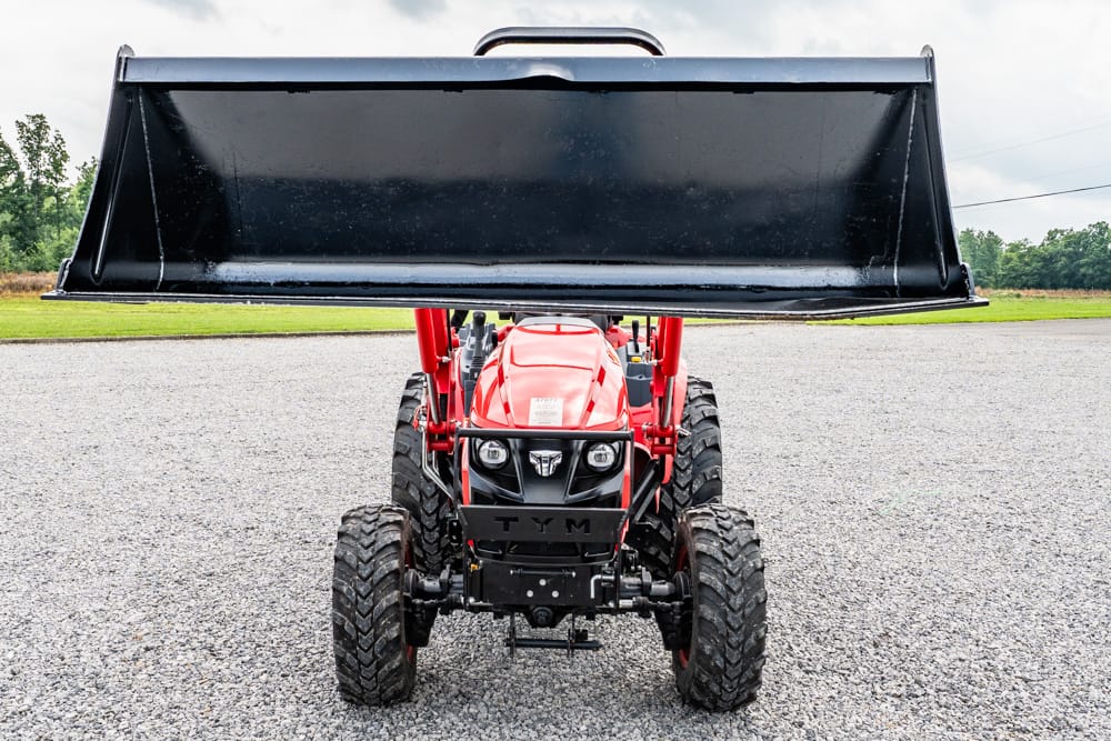 Red compact tractor with a large black front-end loader raised overhead on a gravel surface.
