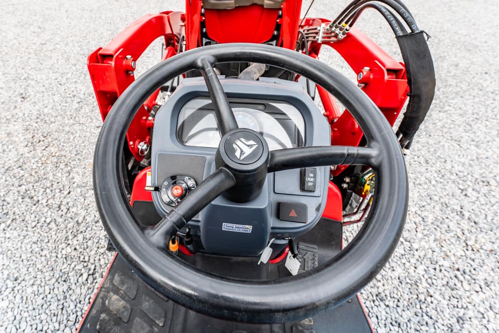 Close-up of a tractor's steering wheel and dashboard with red machinery arms in the background on a gravel surface.