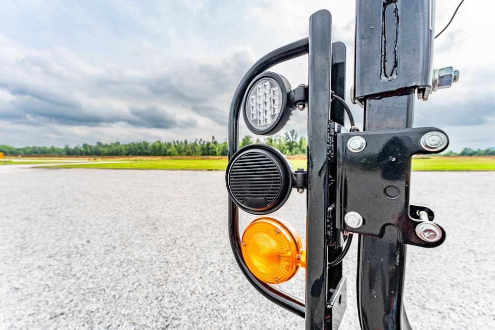 Close-up of a trailer light assembly mounted on a black metal hitch, featuring a white LED light, a black circular vent, and an orange indicator light against a cloudy sky and open lot.