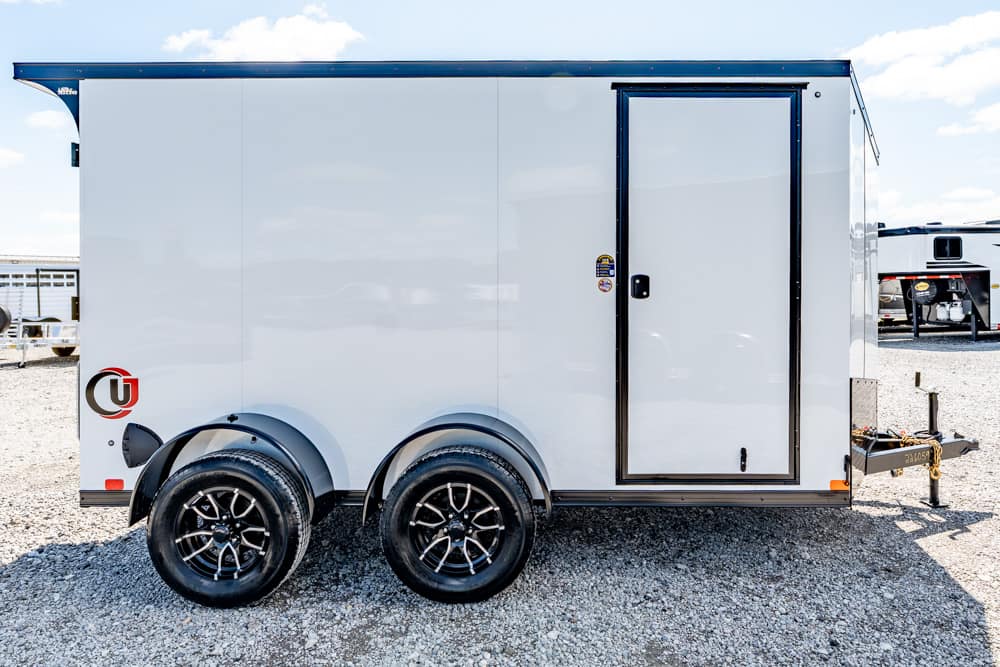 White dual-axle cargo trailer on a gravel lot, side door closed and black-rimmed wheels visible.