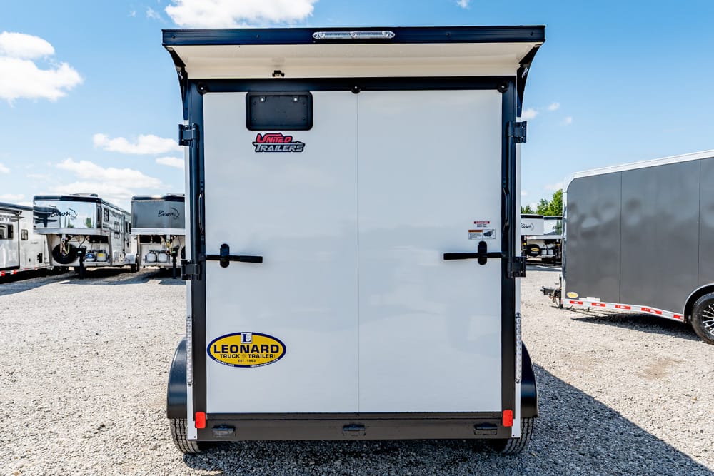Rear view of a white enclosed cargo trailer with double doors, black latches, and Leonard trailer sticker on a gravel lot with other trailers nearby.