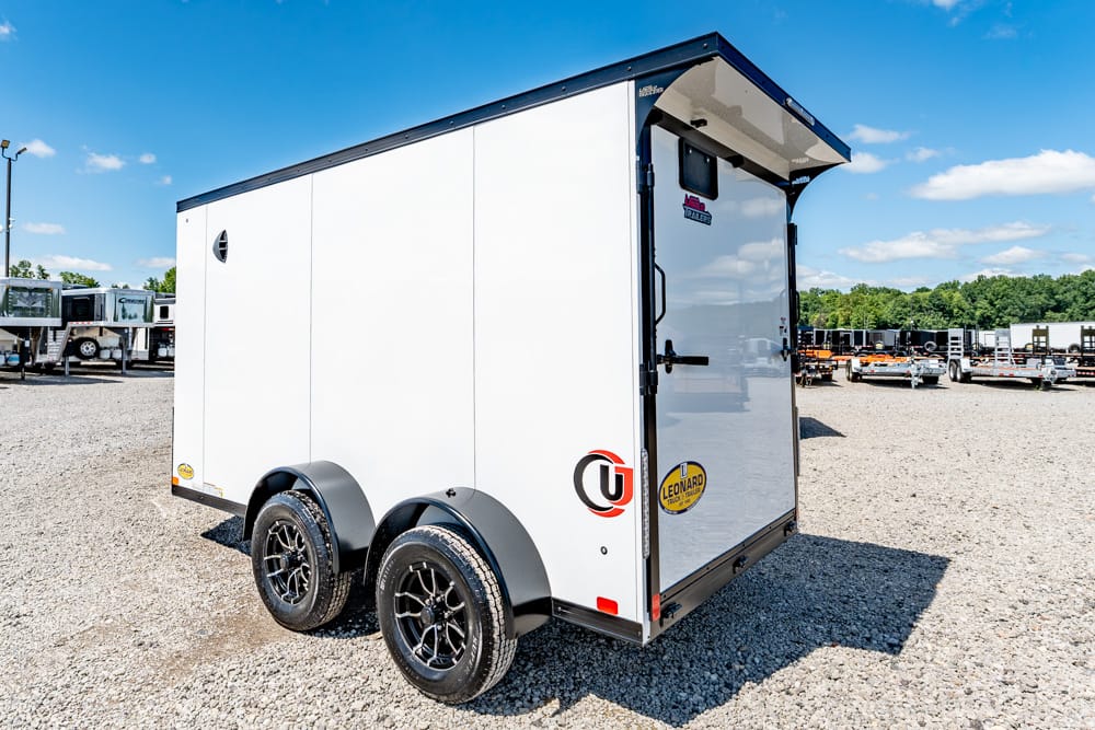 White double-axle enclosed cargo trailer on a gravel lot, with ramp rear and blue sky above
