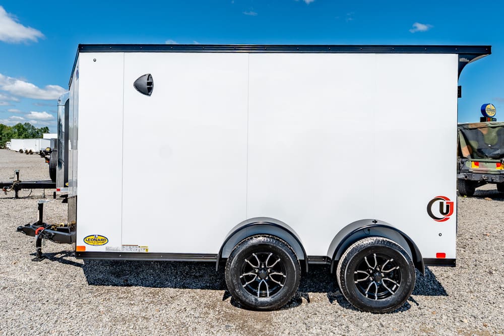White enclosed cargo trailer with dual axles and black rims on a gravel lot under a blue sky.
