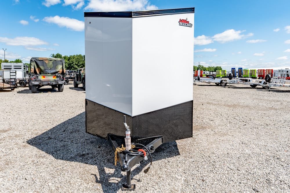 Front view of an enclosed trailer with a white upper section and black diamond-plate lower section, hitch and safety chains in the foreground on a gravel lot.