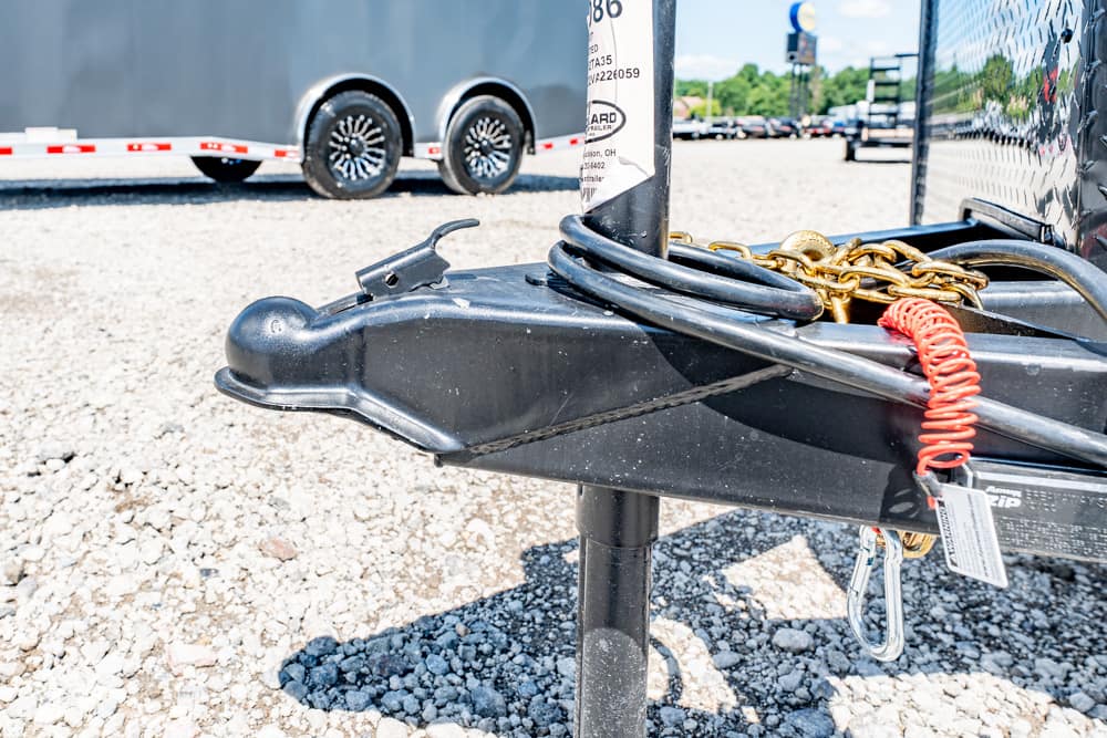 Close-up of a black trailer tongue with a safety chain, red coiled cord, and padlock on a gravel lot.