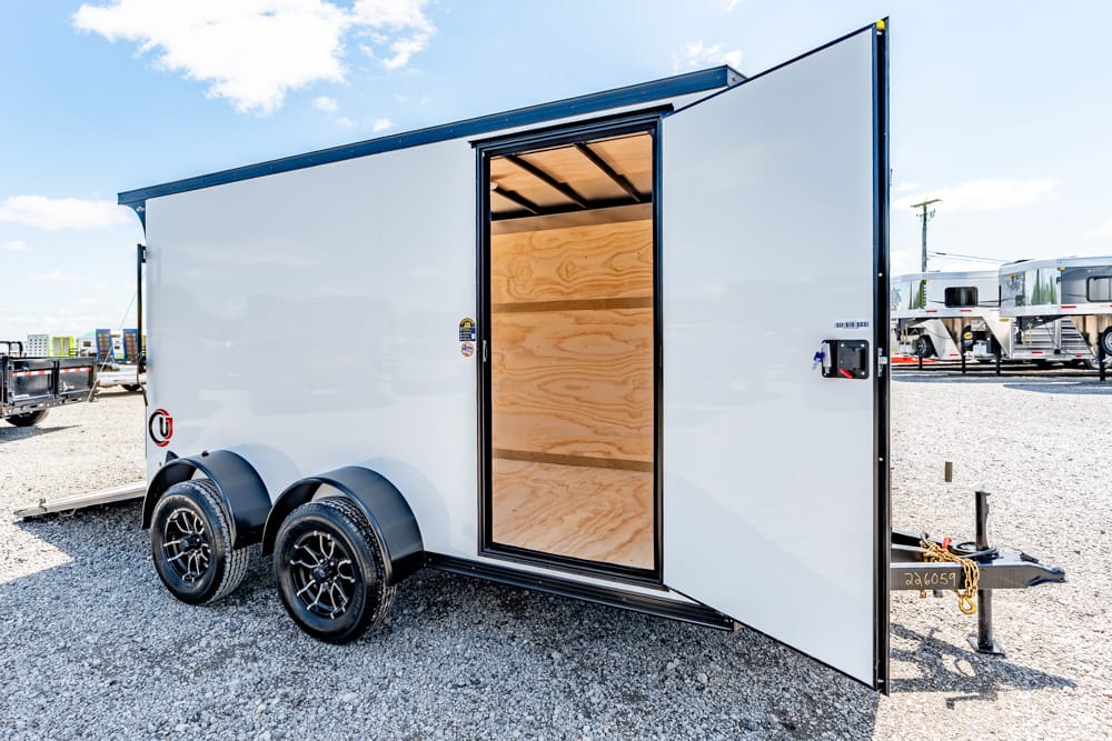 White enclosed cargo trailer with dual axles, open rear door revealing plywood interior on a gravel lot.