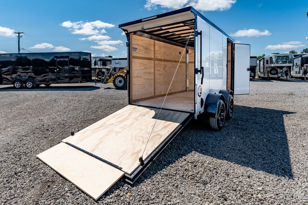 White, dual-axle enclosed trailer with the ramp lowered, empty plywood interior visible on a gravel lot with other trailers in the background.