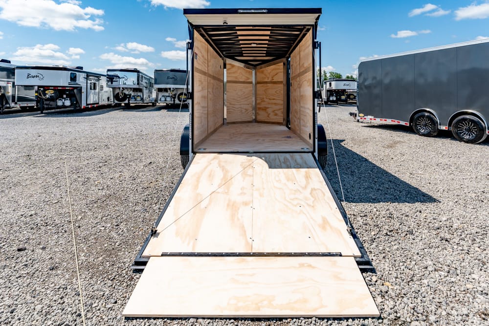 Open cargo trailer with wooden interior, ramp lowered onto a gravel lot, other trailers visible in the background.