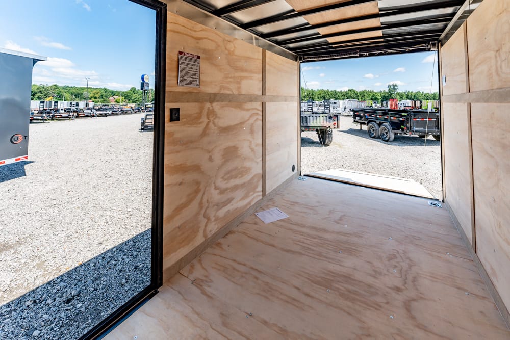 Interior of an empty plywood cargo trailer with rear ramp open to a gravel lot of parked trailers under a blue sky, daytime