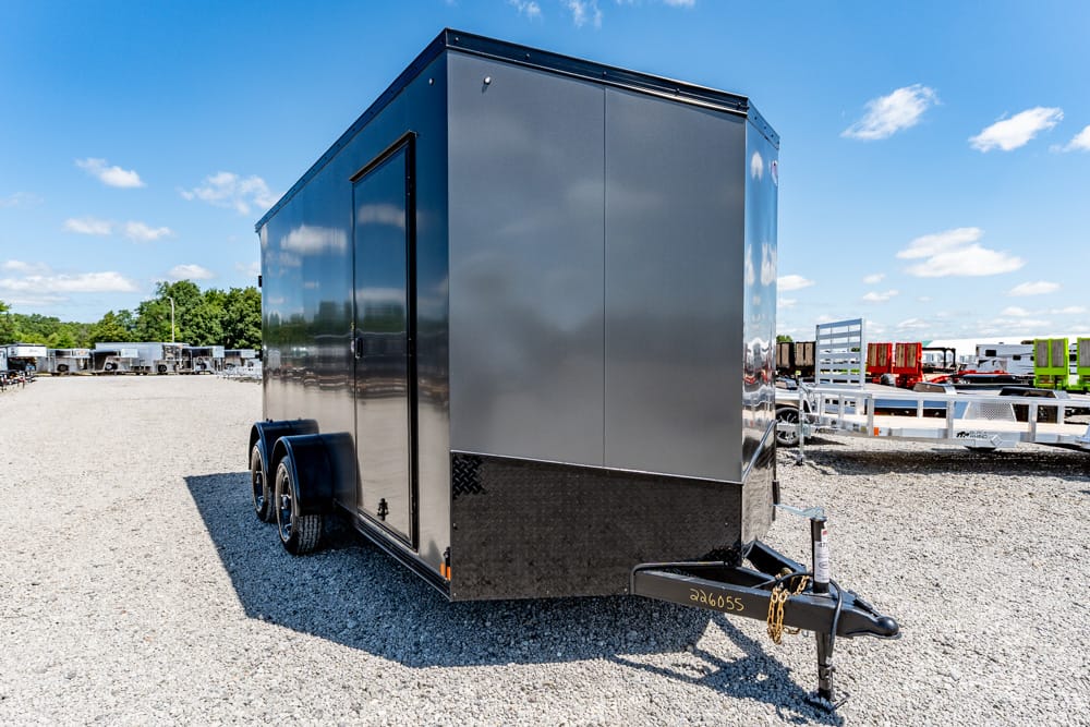 Gray enclosed cargo trailer on a gravel lot with its hitch and jack visible, other trailers in the background under a blue sky.