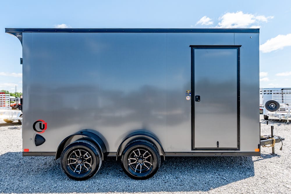 Gray enclosed dual-axle trailer with a side entry door on a gravel lot under a blue sky.