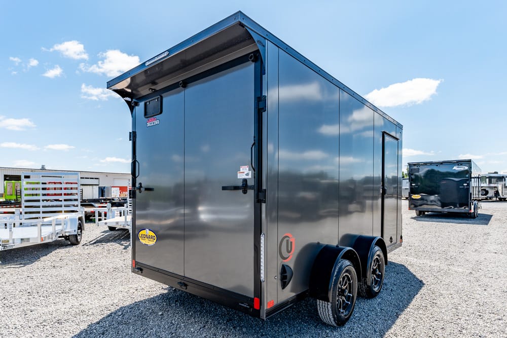 Black enclosed cargo trailer with dual axles and rear doors, parked on a gravel lot with other trailers in the background.