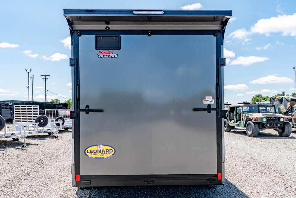 Rear of a gray cargo trailer with Leonard logo, parked on gravel with other trailers and trucks in the background beneath a blue sky.