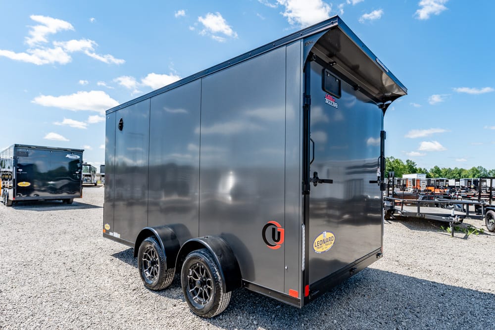 Gray enclosed dual-axle cargo trailer on a gravel lot, side and rear doors closed, with logos on the back panel and clear blue sky above.