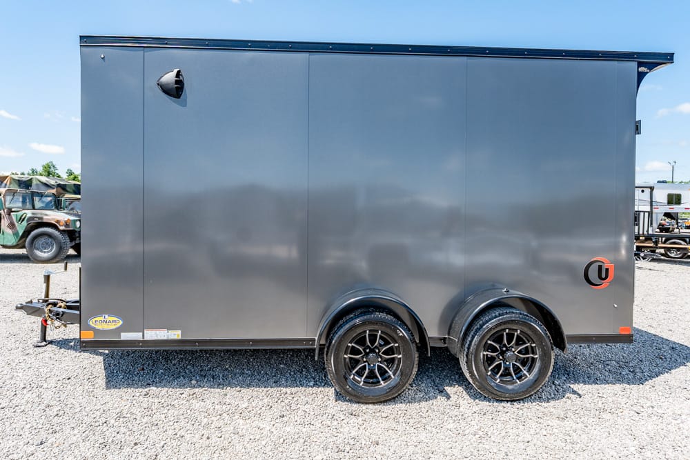 Gray enclosed dual-axle cargo trailer on a gravel lot, with a front hitch and two black alloy wheels visible on the side.”,
