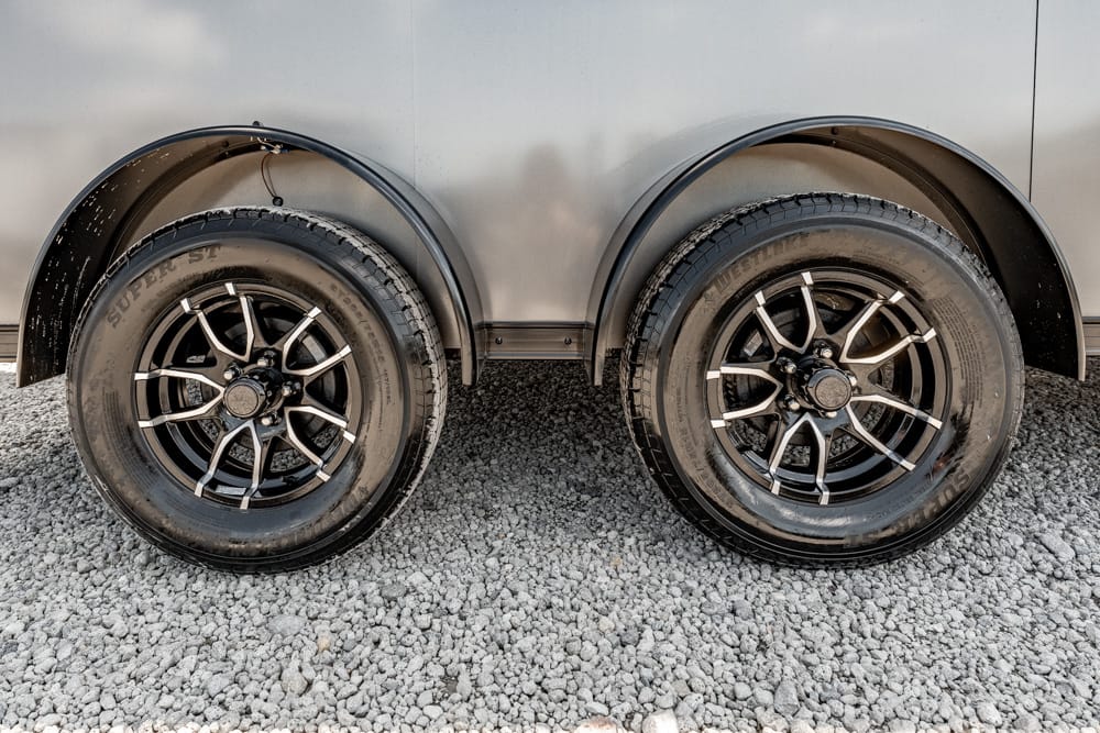 Dual trailer wheels with black and silver rims on a gravel surface, silver fenders overhead.