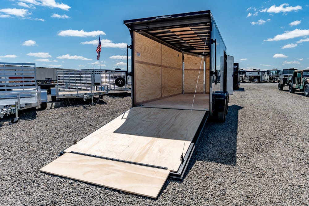 Open black cargo trailer with the rear ramp lowered for loading, showing plywood interior on a gravel lot with other trailers nearby and an American flag in the background.