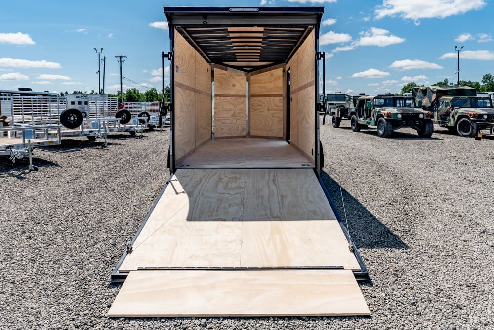 Open empty cargo trailer with wooden interior and ramp lowered on a gravel lot, surrounded by parked trucks and trailers on a sunny day