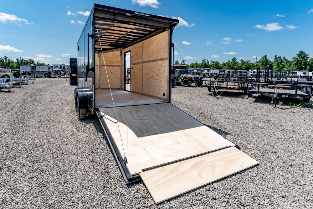 Open cargo trailer with wooden interior and ramp lowered on a gravel lot.