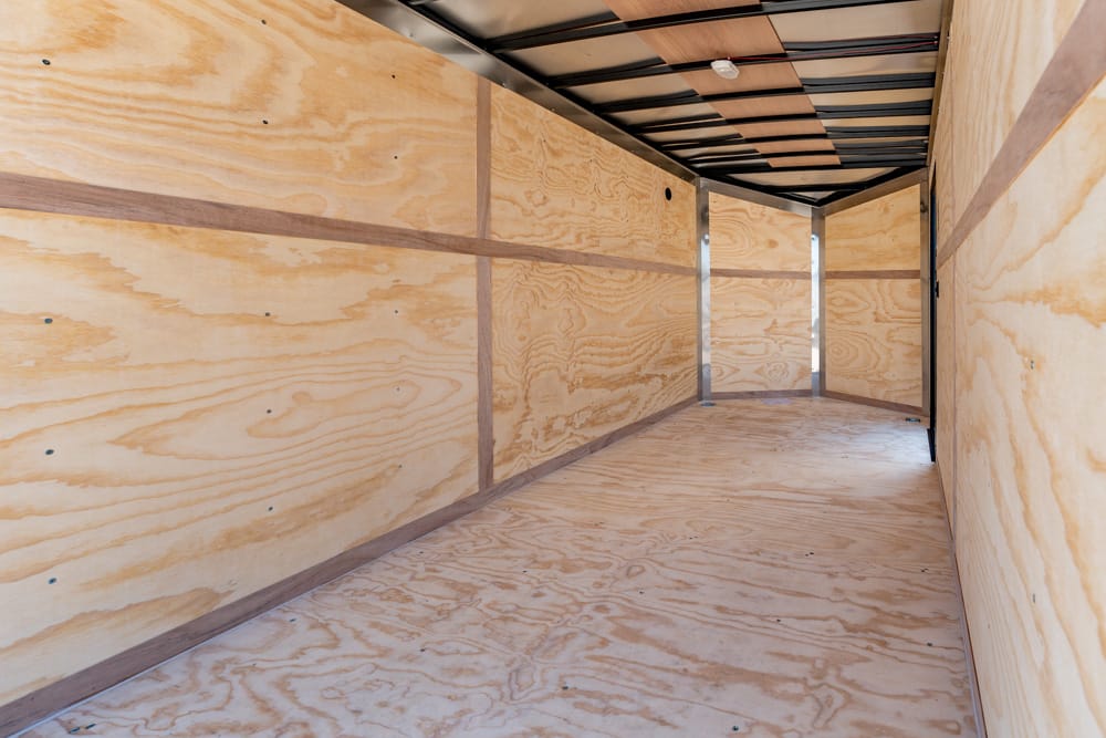 Empty plywood cargo trailer interior with light wood walls, floor, and exposed metal framing along the ceiling and corners.
