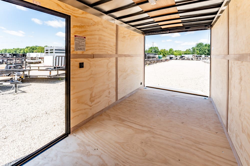 Interior of an empty cargo trailer with plywood walls and floor, open rear door showing a gravel lot with parked trailers under a blue sky.