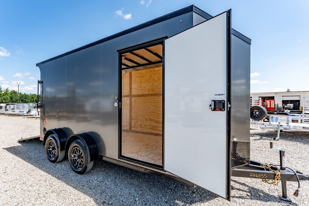 Enclosed dual-axle trailer with open door revealing plywood interior, parked on gravel lot under a blue sky.