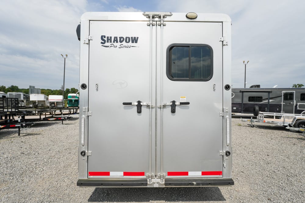 Rear view of a silver Shadow Pro Series trailer with double doors, a small window, and red reflectors on a gravel lot.